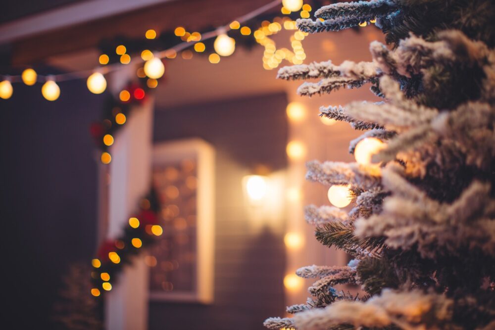 Close up of Christmas snowy fir tree outdoors with house in background. Yard and columns decorated with garlands and string lights.